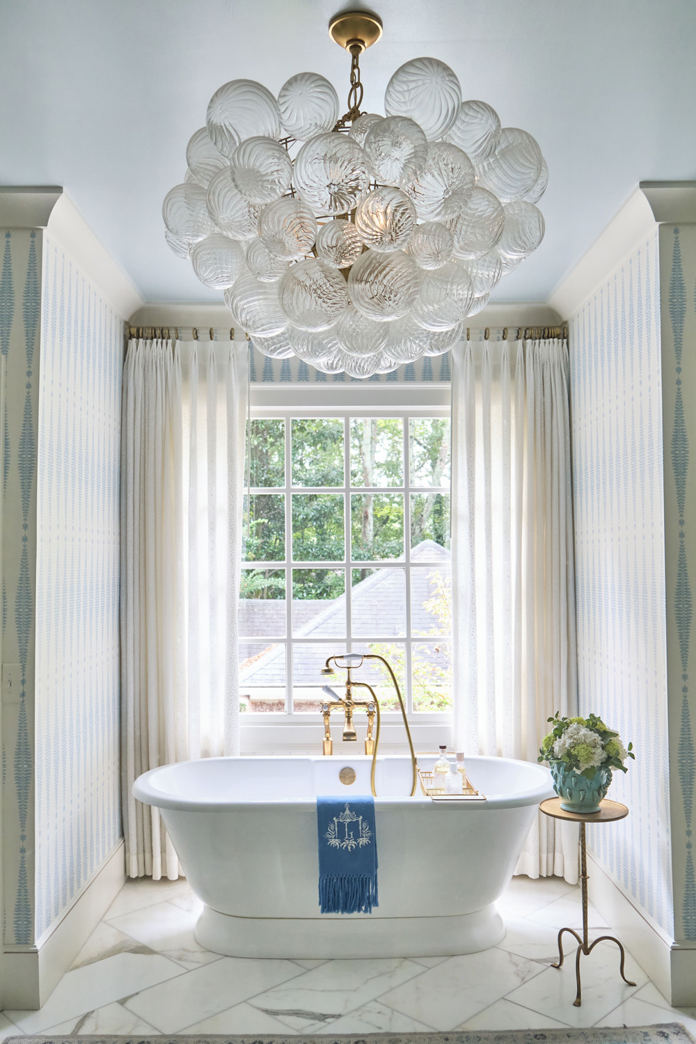 a classic tub with handheld shower sits in front of a large window with blue and white patterned wallpaper